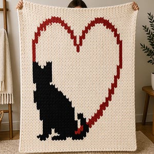 Puede incluir: Manta de punto de color crema con un gran contorno de corazón rojo y una silueta de gato negro. La manta está hecha de hilo grueso, creando una apariencia texturizada. El gato está sentado dentro de la forma de corazón. La manta es sostenida por una persona.
