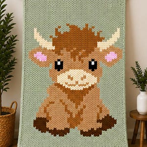 May include: A sage green chunky knit blanket featuring a large, centered graphic of a brown Highland cow. The cow has cream-colored horns, pink inner ears, and dark brown hooves. The blanket is displayed hanging from a wooden rod.