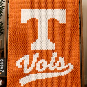 May include: A chunky knit blanket in bright orange featuring a large white letter T and the word Vols in white script lettering. The blanket is constructed with thick yarn in a prominent vertical stitch pattern, creating a textured surface.