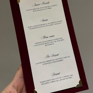 May include: A rectangular menu card with a deep burgundy velvet border and gold-toned metal corner protectors. The white center features black printed text listing courses: Amuse-Bouche, Starter, Main course, Pre-Dessert, and Dessert with descriptions.