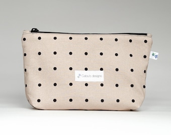 May include: A beige cosmetic bag with a black zipper and black polka dots. The bag has a rectangular shape and a small white tag with the text "Catou's designs". The bag is made of a soft fabric.
