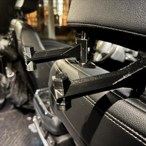 May include: Black car headrest mount with two horizontal arms. The mount is attached to a black leather car seat with white stitching. The mount is designed to hold a tablet or other device.