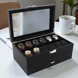 May include: A black watch display case with a glass top, showcasing several wristwatches. The case has a drawer and a silver clasp. A white mug of coffee sits nearby.
