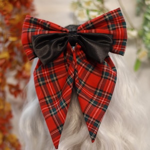 May include: A red tartan bow with a black satin centre. The bow is attached to a wig with long, wavy, grey hair. The tartan pattern includes green and white lines. The bow is a decorative hair accessory.