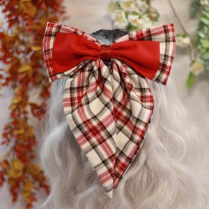 Peut inclure: Un grand nœud rouge avec un motif à carreaux rouge, noir et crème. Le nœud est attaché à une chevelure grise. Le nœud est fait de deux tissus différents, un rouge uni et un tissu à carreaux.