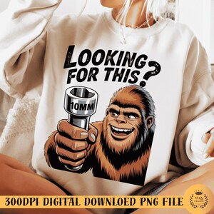 Puede incluir: Sudadera blanca con un diseño gráfico de un Bigfoot sonriente sosteniendo una llave de vaso de 10 mm. El texto superior dice "LOOKING FOR THIS?". Es un archivo PNG de descarga digital.