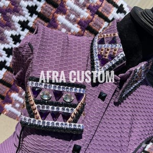 Könnte beinhalten: Nahaufnahme eines lila Reitshirts mit dem Text "AFRA CUSTOM". Das Shirt hat ein strukturiertes Muster und ist mit schwarzen, goldenen und silbernen Strasssteinen und lila Edelsteinen verziert. Kragen und Manschetten sind aufwendig verziert.