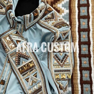 Könnte beinhalten: Ein hellblaues Show-Shirt mit aufwendigen Verzierungen. Das Shirt hat detaillierte Perlenarbeiten und dekorative Akzente in Gold-, Braun- und klaren Kristalltönen. Der Text "AFRA CUSTOM" ist auf der Vorderseite des Shirts zu sehen.