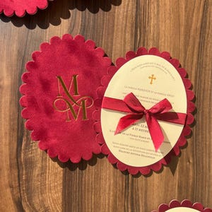 Può includere: Inviti di nozze in velluto bordeaux con iniziali dorate in rilievo e bordi smerlati. Ogni invito è legato con un nastro rosso e include testo stampato con un simbolo a croce.