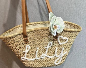 May include: A woven straw tote bag featuring tan leather straps and a white crocheted script reading Lily. The bag is decorated with a white orchid flower accent and a small white crocheted heart shape on the front exterior surface.