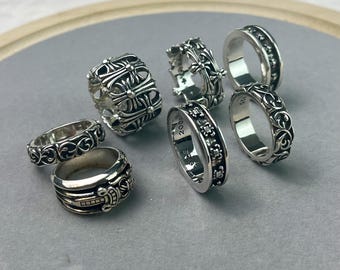 Könnte beinhalten: Eine Sammlung von sechs Silberringen mit komplexen Prägemustern, darunter florale Motive, Kreuzsymbole und ein Dolchdetail. Jeder Ring zeigt polierte Metalloberflächen mit dunklen vertieften Bereichen, die die Gravuren hervorheben.