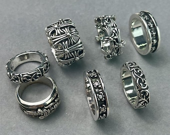 May include: A collection of six silver rings featuring intricate embossed patterns, including cross motifs, floral scrollwork, and geometric designs. The rings vary in width and thickness, showcasing polished metallic surfaces with dark recessed detailing.