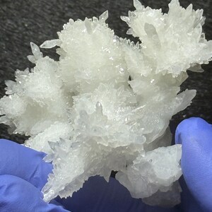 Puede incluir: Un racimo de formaciones minerales cristalinas de color blanco transl&uacute;cido con bordes afilados y puntiagudos. La estructura presenta m&uacute;ltiples puntos de ramificaci&oacute;n y una textura superficial irregular y dentada que refleja la luz, creando un aspecto brillante.
