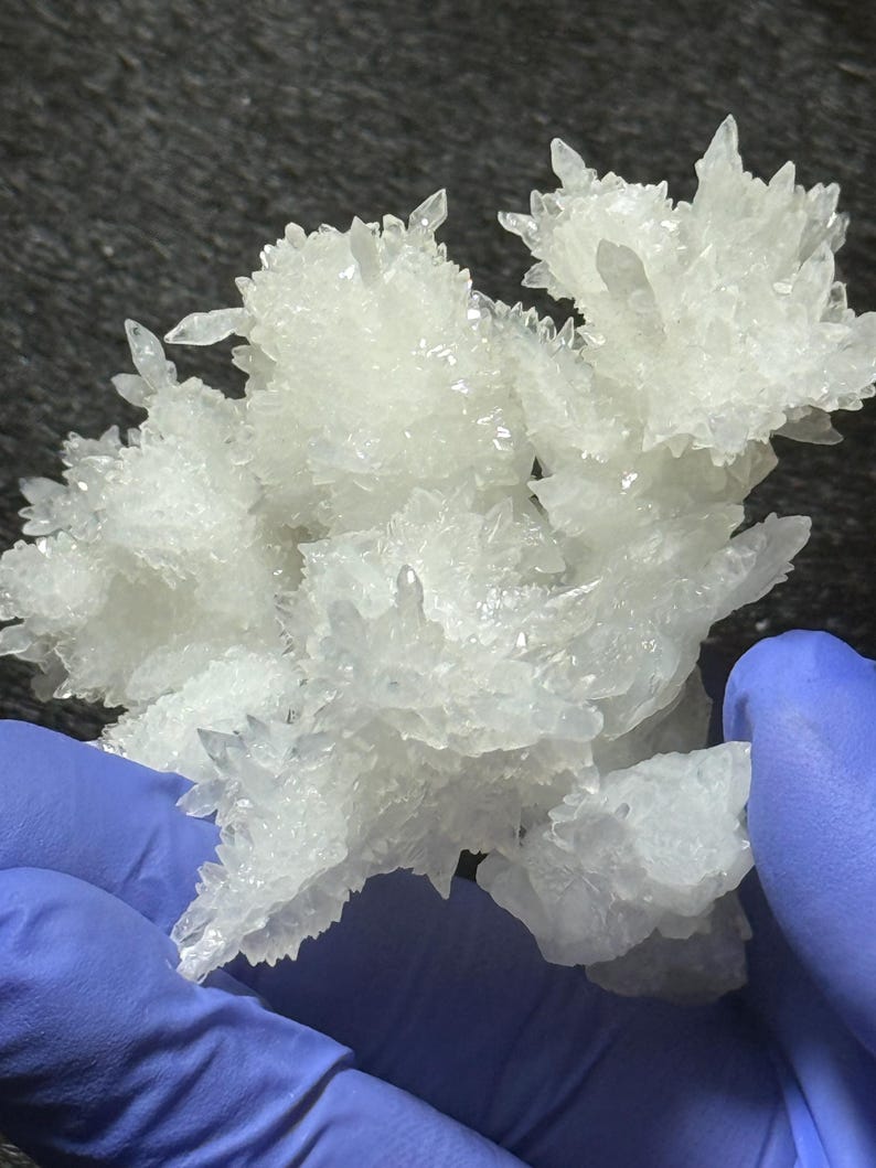 Puede incluir: Un racimo de formaciones minerales cristalinas de color blanco transl&uacute;cido con bordes afilados y puntiagudos. La estructura presenta m&uacute;ltiples puntos de ramificaci&oacute;n y una textura superficial irregular y dentada que refleja la luz, creando un aspecto brillante.