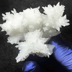 Puede incluir: Un racimo de cristales blancos transl&uacute;cidos con bordes afilados y puntiagudos y una estructura dentada y ramificada. La formaci&oacute;n mineral presenta m&uacute;ltiples puntas radiantes que crean una apariencia compleja y multifac&eacute;tica con una superficie brillante.