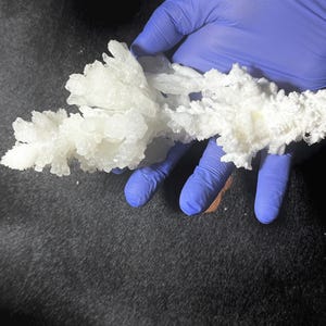 Pu&ograve; includere: Un gruppo di formazioni cristalline bianche, simili a un campione minerale naturale, &egrave; tenuto da una mano guantata. I cristalli hanno una superficie irregolare e strutturata e sono disposti in un motivo ramificato. Lo sfondo &egrave; una superficie scura e strutturata.