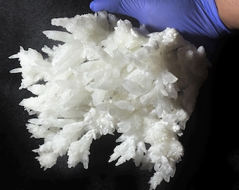 Puede incluir: Un grupo de cristales blancos con formaciones afiladas y puntiagudas. Los cristales tienen un aspecto esmerilado y son sostenidos por una mano enguantada. La forma general es irregular y texturizada, con una estética natural y orgánica.