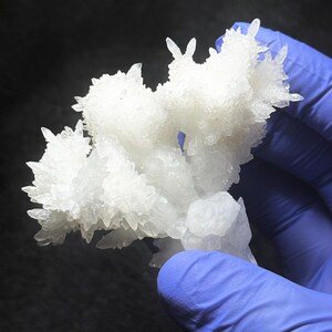 Puede incluir: Primer plano de un grupo de formaciones cristalinas blancas, similar a una geoda o esp&eacute;cimen mineral. Los cristales tienen una textura rugosa y son sostenidos por una mano enguantada sobre un fondo oscuro. Esp&eacute;cimen geol&oacute;gico natural.