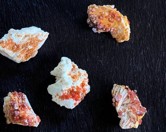 Puede incluir: Cinco especímenes minerales con una mezcla de colores. Los minerales tienen una base blanca con formaciones de cristales naranjas y rojas. El fondo es una superficie oscura y texturizada.