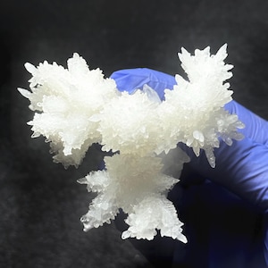 Puede incluir: Primer plano de una formaci&oacute;n cristalina blanca, con m&uacute;ltiples puntas. Los cristales tienen un aspecto esmerilado y una superficie texturizada. El fondo es oscuro, contrastando con los cristales brillantes. La estructura es sostenida por una mano enguantada.