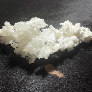 Pu&ograve; includere: Un gruppo di formazioni cristalline bianche, possibilmente un campione minerale, poggia su una superficie scura e strutturata. I cristalli hanno una consistenza ruvida e irregolare e appaiono traslucidi. Un piccolo oggetto marrone &egrave; vicino.