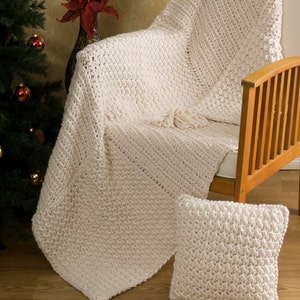May include: A white crocheted throw blanket with a fringe on the bottom edge. The blanket is draped over a wooden chair with a white cushion. A smaller white crocheted pillow is on the floor in front of the chair.