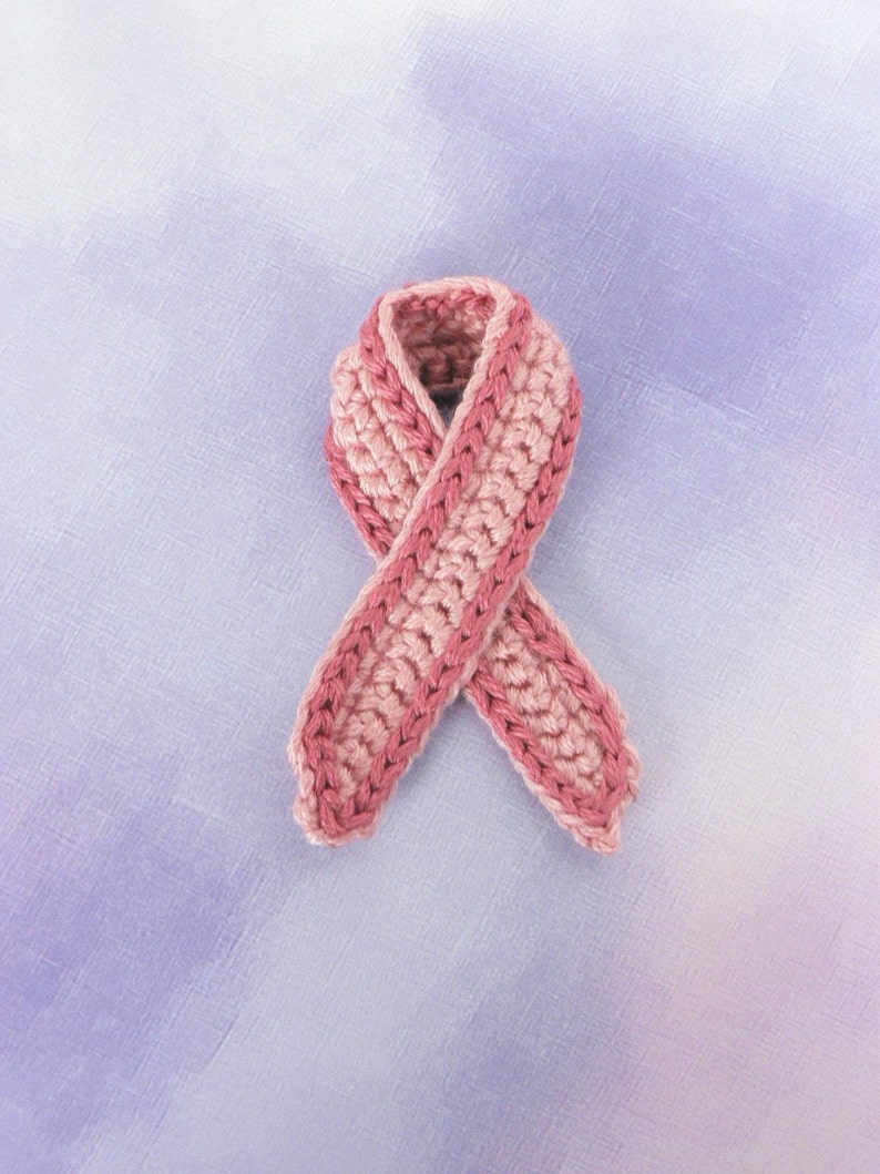 May include: A pink crocheted ribbon tied in a bow. The ribbon is against a light purple background.