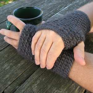 May include: Dark grey knitted fingerless gloves. The gloves are made of a textured yarn and cover the hand up to the knuckles, leaving the fingers free. The gloves are shown on a wooden table.