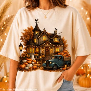 May include: A cream-coloured t-shirt featuring a graphic of a haunted house with glowing windows, a blue pickup truck filled with pumpkins, a ghost, and autumn foliage. The truck bed has the word HARVEST printed on the side of the wooden crate.