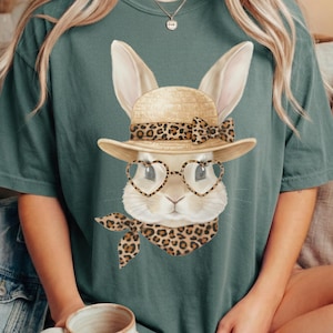 May include: A teal green t-shirt with a bunny graphic. The bunny wears a straw hat with a leopard print band and bow, heart-shaped glasses, and a leopard print scarf. The bunny has long ears and a white face.