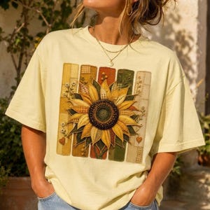Puede incluir: Camiseta amarillo claro con un diseño de girasol. El girasol está centrado en un fondo de retazos de varios colores y patrones. La camiseta tiene mangas cortas y un corte holgado, adecuada para uso casual.