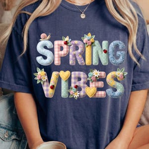 Könnte beinhalten: Marineblaues T-Shirt mit den Worten "SPRING VIBES" in bunten, strukturierten Buchstaben. Die Buchstaben sind mit floralen und Insekten-Akzenten verziert, darunter eine Schnecke und Marienkäfer. Das Shirt hat eine lockere Passform.