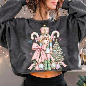 May include: A dark gray long-sleeve sweatshirt featuring a central graphic of a nutcracker soldier wearing a pink and white uniform, accented by a large pink gingham bow, candy canes, a decorated Christmas tree, and various ornaments.