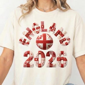 May include: Cream-coloured t-shirt with the word "ENGLAND" above a football graphic, and the year "2026" below. The text and ball feature a red and white patchwork design. The shirt has short sleeves and a round neckline.