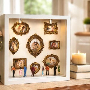 Peut inclure: Un coffret blanc avec plusieurs petits cadres dorés ornés contenant des photos. De petites figurines sont placées en dessous des cadres. Le coffret est éclairé par le haut, et une bougie est posée sur une pile de livres.