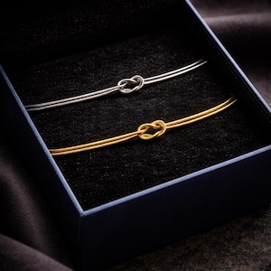 Peut inclure: Deux bracelets à nœud, un argenté et un doré, sont présentés dans un coffret bleu foncé à intérieur noir. Les bracelets sont fins et présentent un motif de nœud au centre.