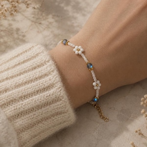 May include: A delicate beaded bracelet featuring white daisy-shaped beads, small white beads, and iridescent blue beads. The bracelet is accented with gold-coloured metal and is shown on a wrist.
