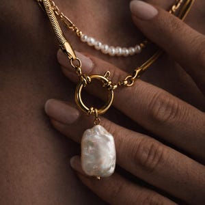 May include: A gold necklace with a large, irregular pearl pendant. The necklace has a circular clasp and is layered with a delicate pearl strand and a chain. The jewellery is displayed against a neutral background.