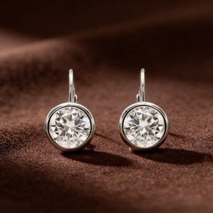 May include: A pair of silver-toned leverback earrings. Each earring features a large, round, clear gemstone set in a bezel setting. The gemstones sparkle, reflecting light. The earrings are displayed against a brown background.