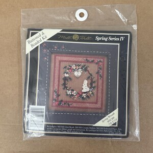 Può includere: Un kit per ricamo Mill Hill Spring Series IV Buttoned & Beaded in un sacchetto di plastica trasparente. La confezione presenta un disegno a punto croce di una ghirlanda con un coniglio bianco e un cuore, incluse perline e bottoni in ceramica.