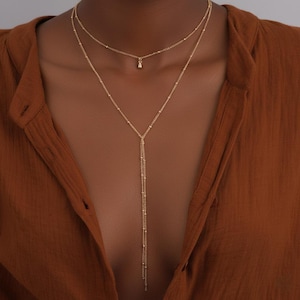 Può includere: Una collana dorata a più strati con una catena delicata e un piccolo ciondolo. La collana presenta un design a forma di Y con una catena più lunga. Il gioiello è presentato su uno sfondo marrone.