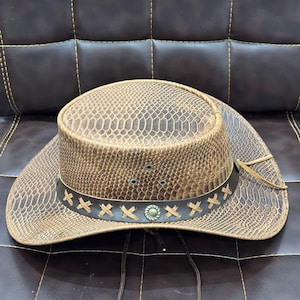 K&ouml;nnte beinhalten: Ein brauner Cowboyhut mit Schlangenmuster. Der Hut hat ein dekoratives Band mit kreuzf&ouml;rmigen N&auml;hten und einer silberfarbenen Verzierung. Ein Kinnriemen ist befestigt. Der Hut liegt auf einer braunen Lederoberfl&auml;che.