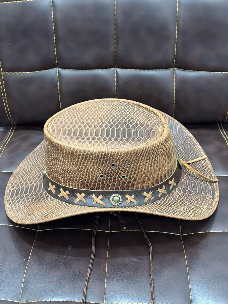 K&ouml;nnte beinhalten: Ein brauner Cowboyhut mit Schlangenmuster. Der Hut hat ein dekoratives Band mit kreuzf&ouml;rmigen N&auml;hten und einer silberfarbenen Verzierung. Ein Kinnriemen ist befestigt. Der Hut liegt auf einer braunen Lederoberfl&auml;che.