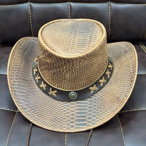 K&ouml;nnte beinhalten: Brauner Cowboyhut mit Schlangenmuster. Der Hut hat eine breite Krempe und ein dekoratives Band mit gen&auml;hten Akzenten und Metallnieten. Ein Kinnriemen ist befestigt.