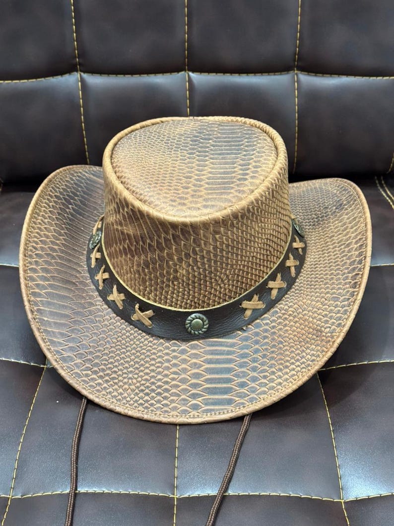 K&ouml;nnte beinhalten: Brauner Cowboyhut mit Schlangenmuster. Der Hut hat eine breite Krempe und ein dekoratives Band mit gen&auml;hten Akzenten und Metallnieten. Ein Kinnriemen ist befestigt.