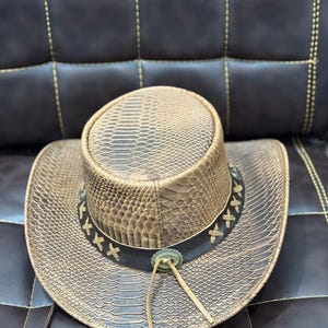 K&ouml;nnte beinhalten: Ein brauner Cowboyhut mit Schlangenmuster. Der Hut hat ein dunkelbraunes Lederband mit dekorativen N&auml;hten und einem silberfarbenen Schmuck. Eine Lederschnur h&auml;ngt vom Hut.