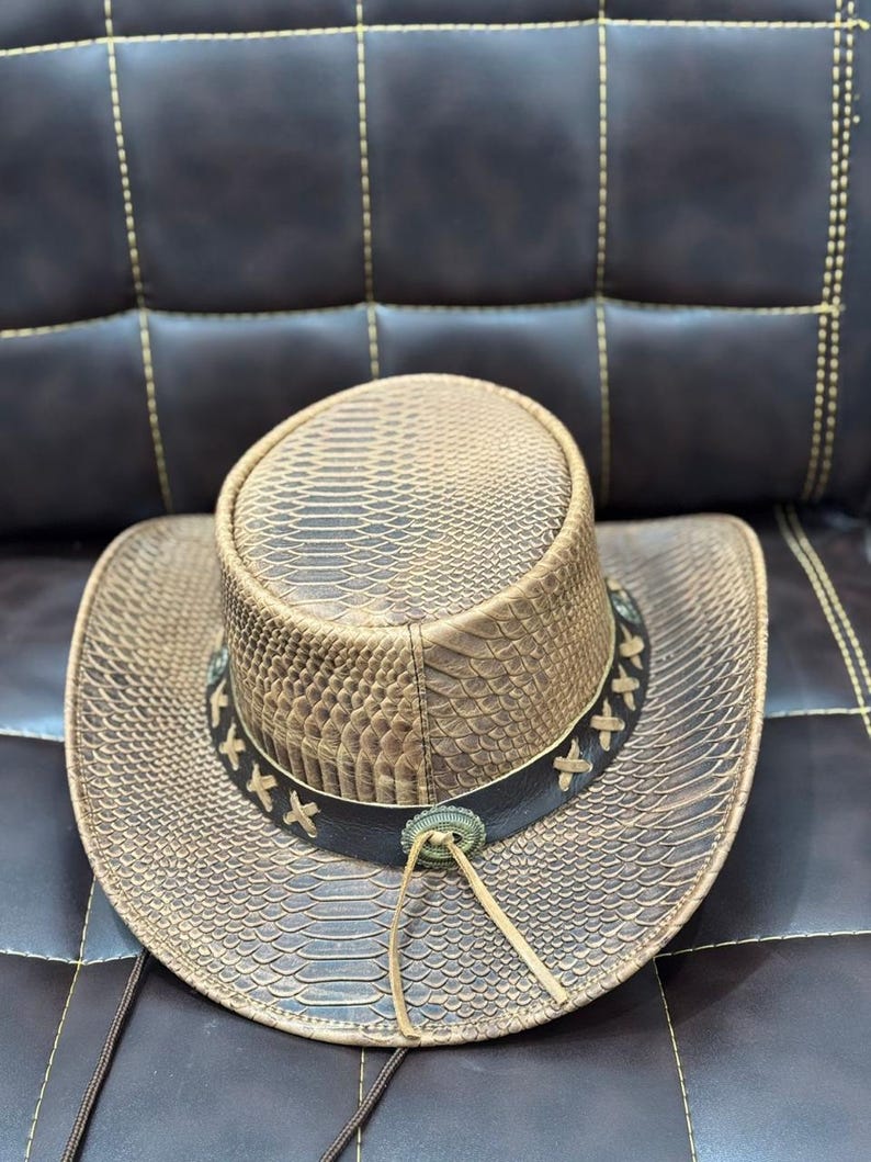 K&ouml;nnte beinhalten: Ein brauner Cowboyhut mit Schlangenmuster. Der Hut hat ein dunkelbraunes Lederband mit dekorativen N&auml;hten und einem silberfarbenen Schmuck. Eine Lederschnur h&auml;ngt vom Hut.