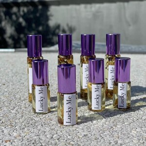 May include: A collection of eight small glass bottles with purple caps. Each bottle contains a clear liquid with visible botanical elements and a label that reads "Lucky Me". The bottles are arranged on a textured surface.