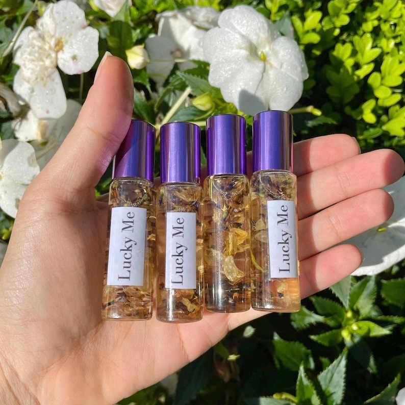 May include: Four small glass roll-on bottles with purple caps are held in a hand. Each bottle contains a clear liquid with dried botanicals and a label that reads "Lucky Me". The background features white flowers and green foliage.