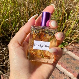 May include: A clear, rectangular perfume bottle with a purple cap and the words "Lucky Me" on a white label. The bottle contains dried flowers and herbs. The bottle is held in a hand against a background of green foliage.
